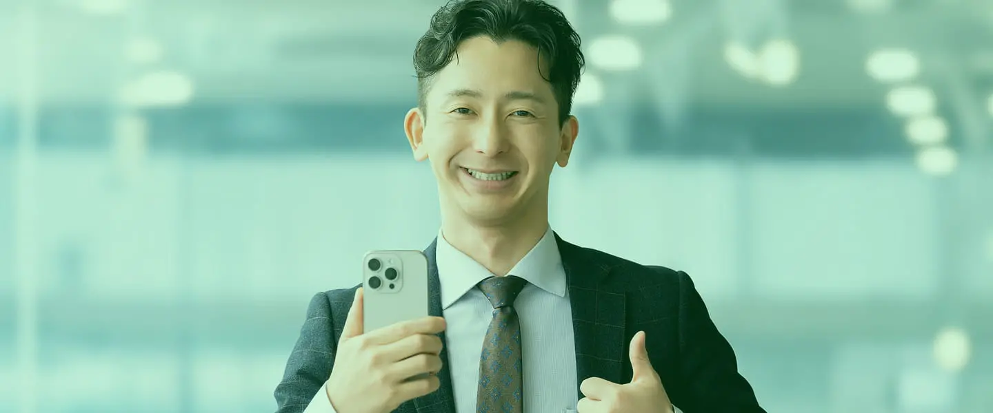 スーツ姿の男性がスマートフォンを手に笑顔で親指を立てるポーズ、明るい室内背景