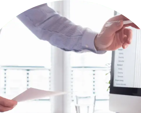 ディスプレイを指しながら資料を持つ人物の手元、オフィスでのプレゼン風景