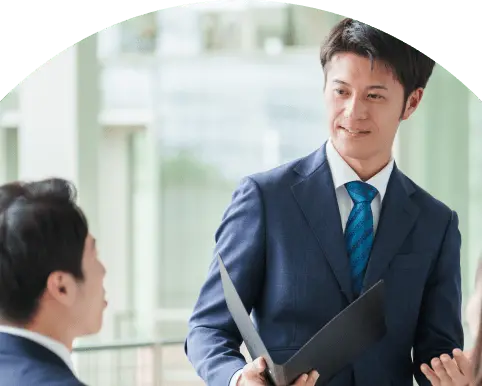 スーツ姿の男性が資料を手にしながら座っている人物に説明をしているビジネスシーン