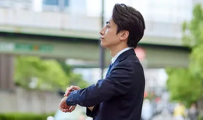 スーツ姿の男性が屋外で腕時計を見ている様子。背景には高架や都市の風景が写る