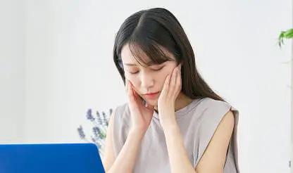 ノートパソコンの前で両手を頬に当て、悩んだ表情を浮かべる女性の室内シーン