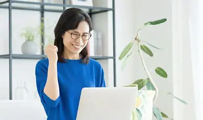 ノートパソコンを見ながらガッツポーズを取る笑顔の女性。明るい部屋で仕事中の様子